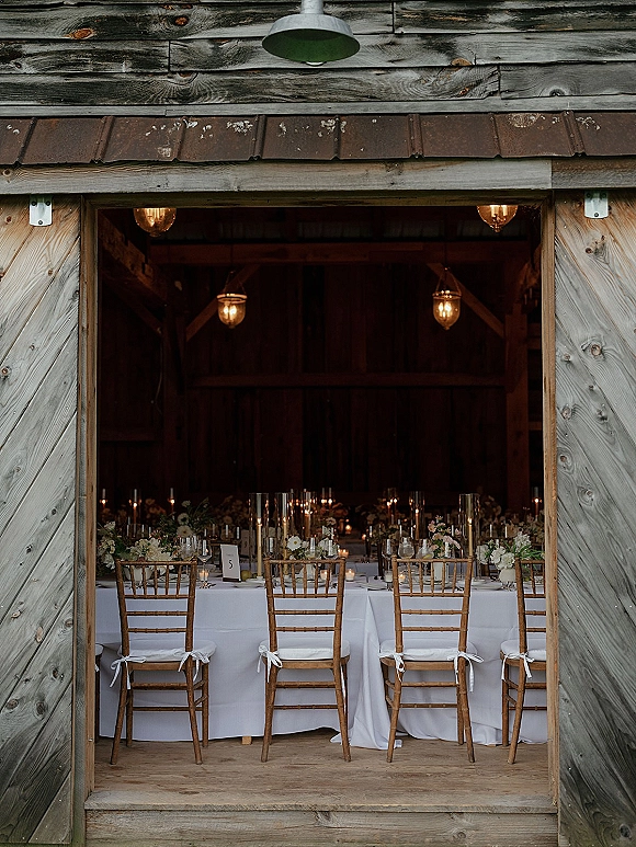 Barn reception tablescape with long banquet tables in white linens, chiavari chairs with ribbons, floral centerpieces and candles in a rustic barn interior