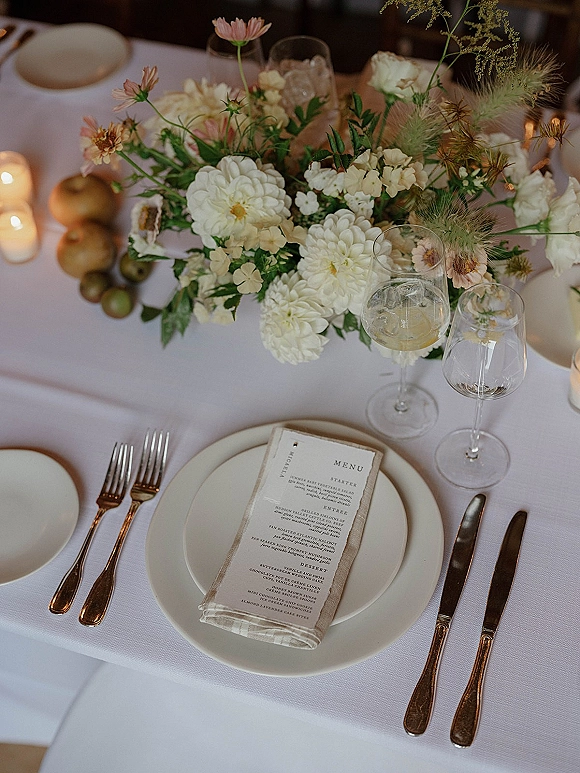 Reception tablescape with wedding table centerpiece of white flowers and greenery, taper and votive candles, menu card, wine glasses on white linens
