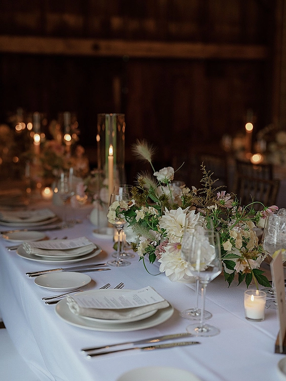 Reception tablescape with wedding table centerpiece, cylinder and votive candles, white linens, plates, menus and glassware in a dim room