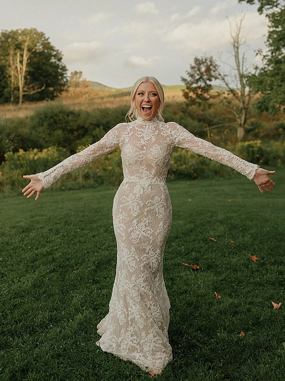 Bridal portrait of a bride in a long sleeve lace wedding dress with arms outstretched on a grassy lawn with hills under cloudy sky