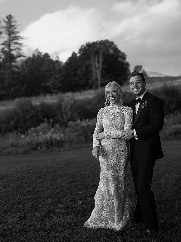 Couple portrait in a black and white wedding portrait style, bride in long sleeve lace gown and groom in tuxedo on a meadow under cloudy sky