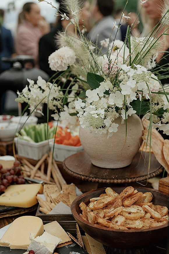 Wedding grazing table with wedding charcuterie table spread of cheese, crackers, bread and grapes, plus a floral centerpiece in a ceramic vase at reception