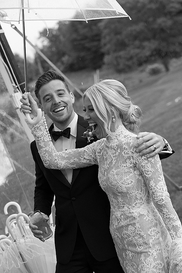 Wedding couple portrait of bride and groom under umbrella, laughing in the rain on a bridge, her lace long sleeves and pearl earrings visible
