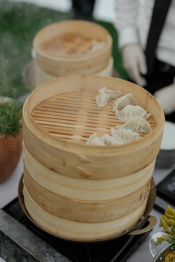 Wedding dumpling station with bamboo steamer baskets of steamed dumplings on a linen buffet table, portable burner, greenery, and servers nearby