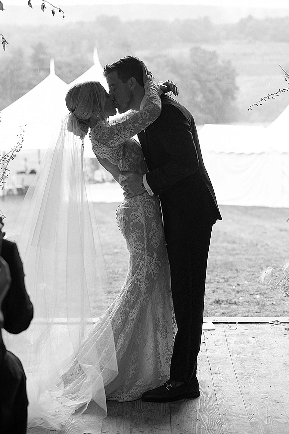 Wedding kiss portrait of bride and groom kissing, her lace dress and long veil flowing on a wooden platform with tents and hills behind
