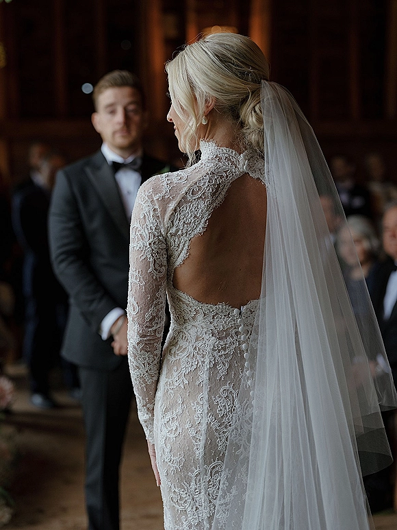 Ceremony moment as bride from behind in lace long sleeve gown and veil walks toward groom in tuxedo at warm wood indoor venue with guests