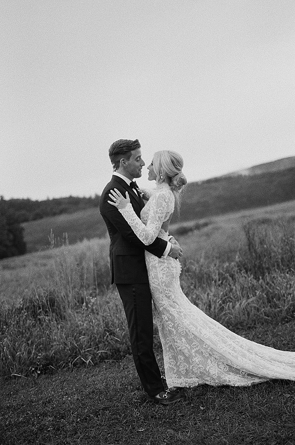 Wedding couple portrait of bride and groom embracing, forehead touch, her lace long-sleeve gown and train in a grassy field with rolling hills