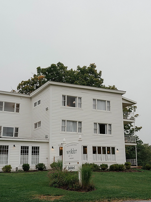 Wedding venue exterior of a white clapboard venue with porch railings and venue sign, framed by shrubs and ornamental grasses under overcast sky