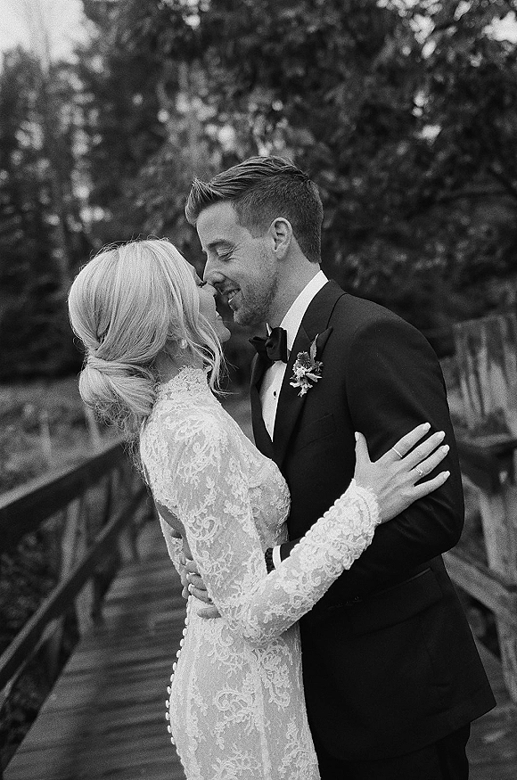 Wedding kiss portrait of bride and groom kissing on a wooden bridge, her long-sleeve lace gown against his tuxedo and bow tie