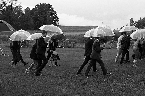 Wedding guests with umbrellas walk across a grassy field under a cloudy sky, wearing suits and cocktail dresses with clear umbrellas