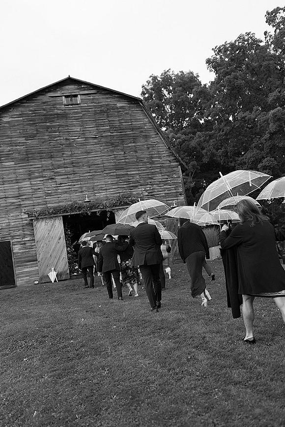 Wedding guests arriving with umbrellas, including a clear umbrella, walking across a lawn toward a rustic barn under an overcast sky