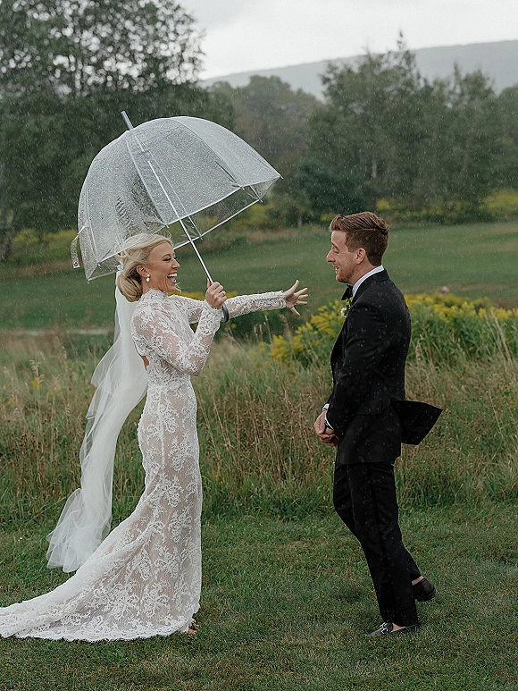 Wedding first look as bride in long lace dress and veil runs to groom under a clear umbrella in rain, in a wildflower field