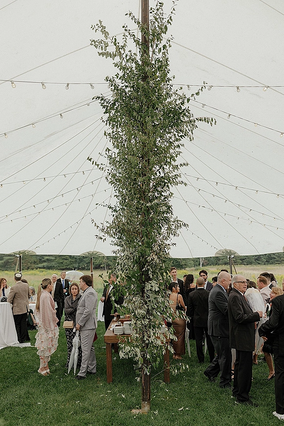 Wedding cocktail hour under a tent with string lights, guests mingling by linen cocktail tables and a wooden buffet on a grassy lawn