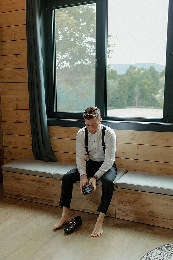 Groom getting ready, putting on dress shoes in black suspenders by a large window with mountains and trees beyond the wood-paneled room