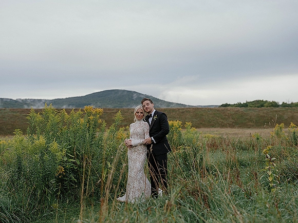 Couple portrait in a wildflower meadow, bride in lace long-sleeve dress and groom in tuxedo with boutonniere by a cloudy mountain ridge