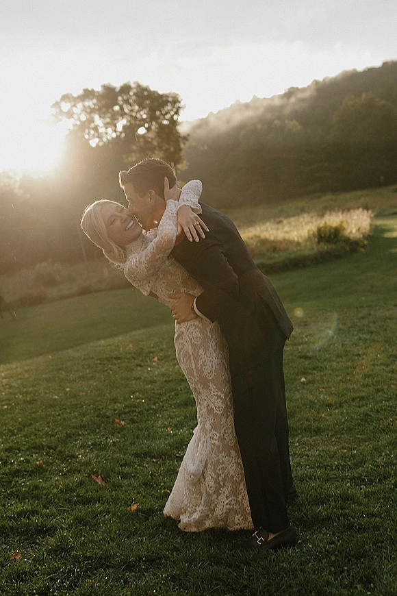 Couple portrait at sunset with groom kissing bride’s cheek as she laughs in a long-sleeve lace wedding dress in a grassy field