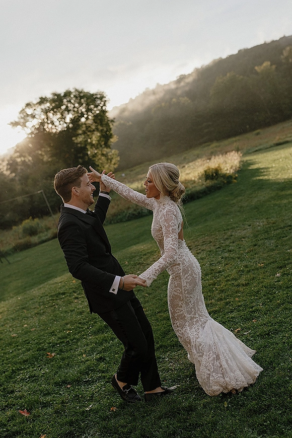 Couple portrait of bride in long sleeve lace wedding dress and groom in tuxedo twirling in a grassy field at sunset with rolling hills