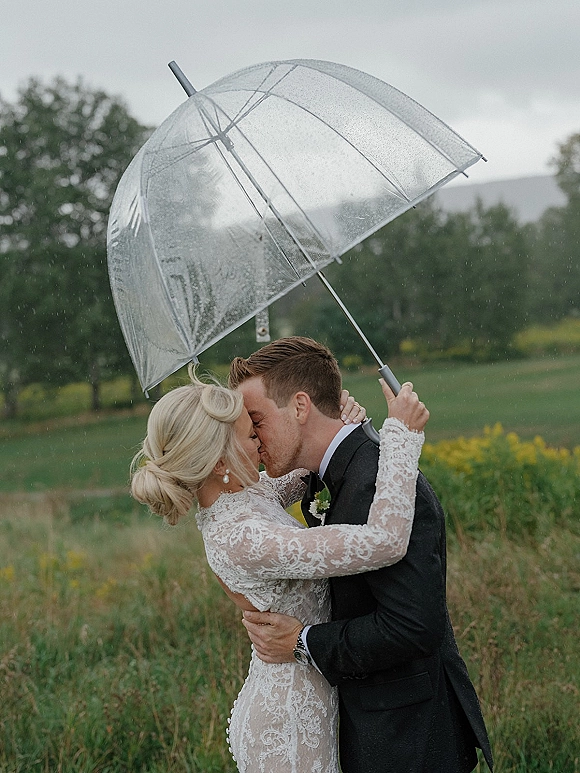 Wedding kiss portrait of couple kissing under umbrella, bride in lace long sleeves and groom in tuxedo amid rainy meadow and trees