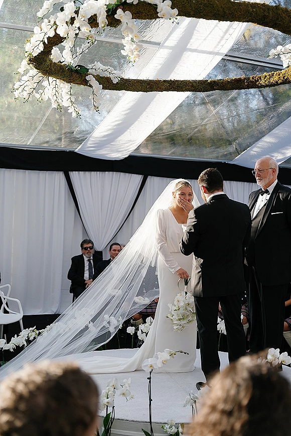 Wedding vows as bride in a white dress covers her mouth, holding an orchid bouquet beside groom in tux under a clear top tent