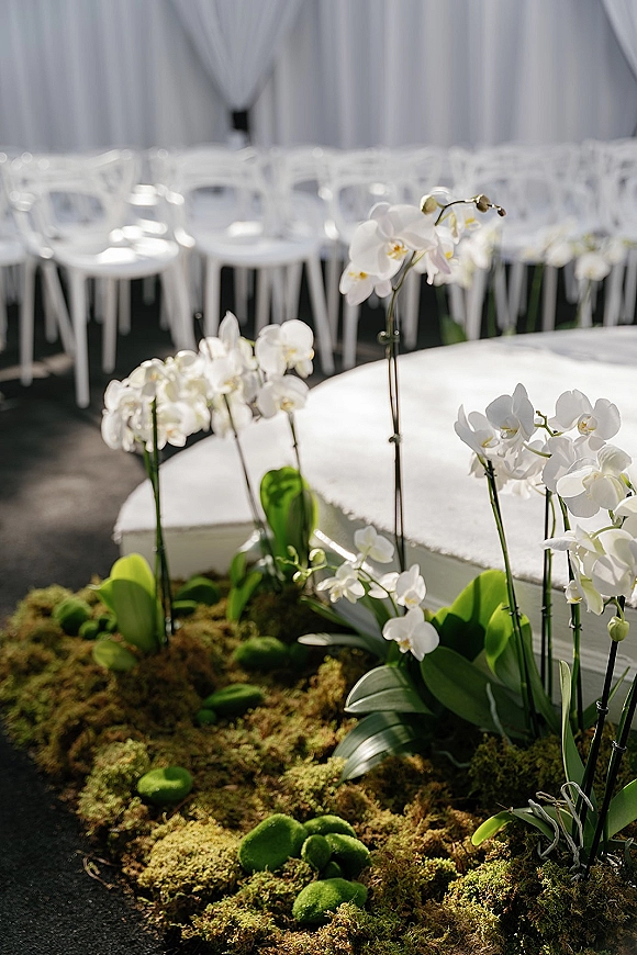 Ceremony aisle decor with orchid aisle flowers and moss greenery lining a white platform, leading to white draping in an indoor venue