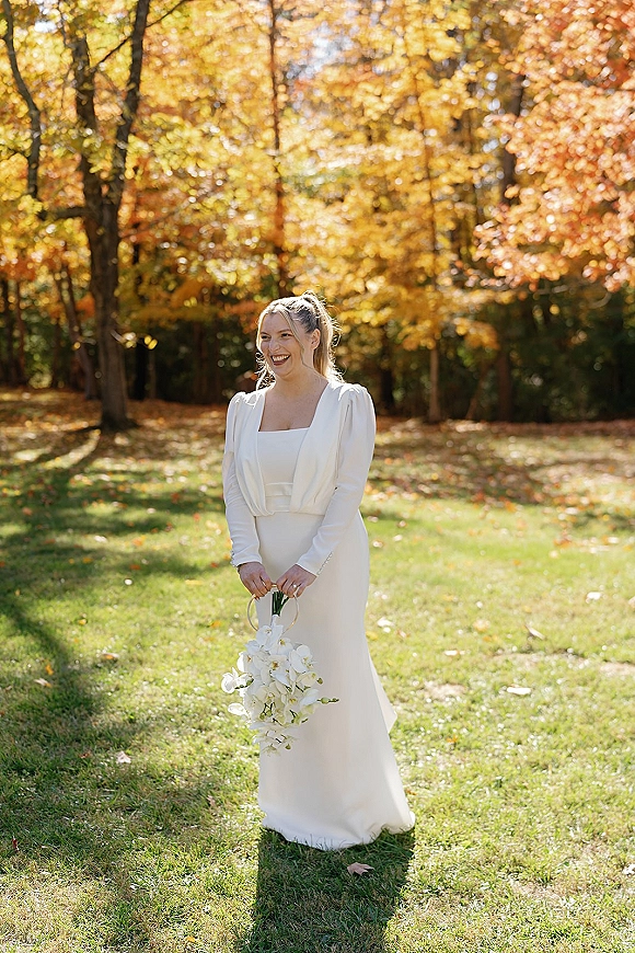 Bridal portrait of a bride laughing in a long-sleeve square-neck dress, holding a cascading orchid bouquet in a sunlit fall park