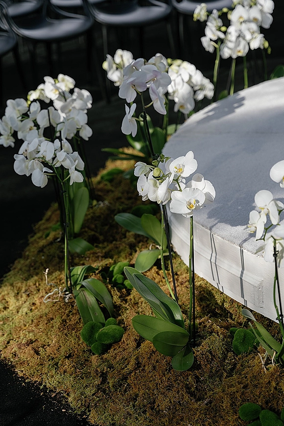 Ceremony aisle decor with orchid aisle flowers on a moss runner, flanked by black chairs and greenery, leading to a white stage in sunlit shadows