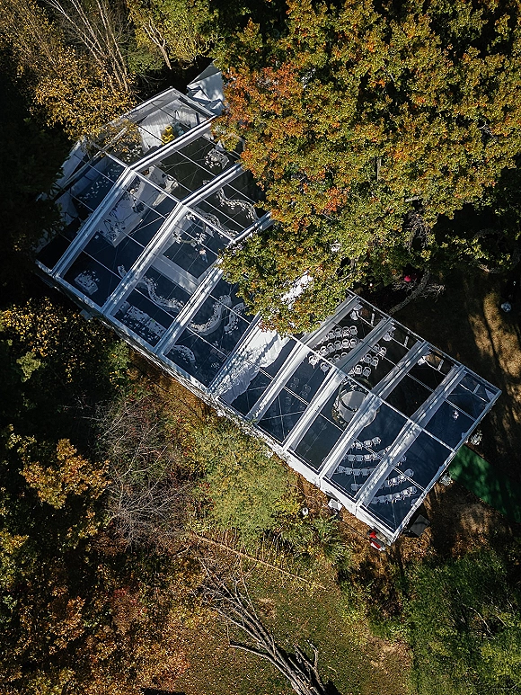 Clear tent wedding with round tables and white chairs under string lights, set on a lawn beside autumn trees in a forest setting