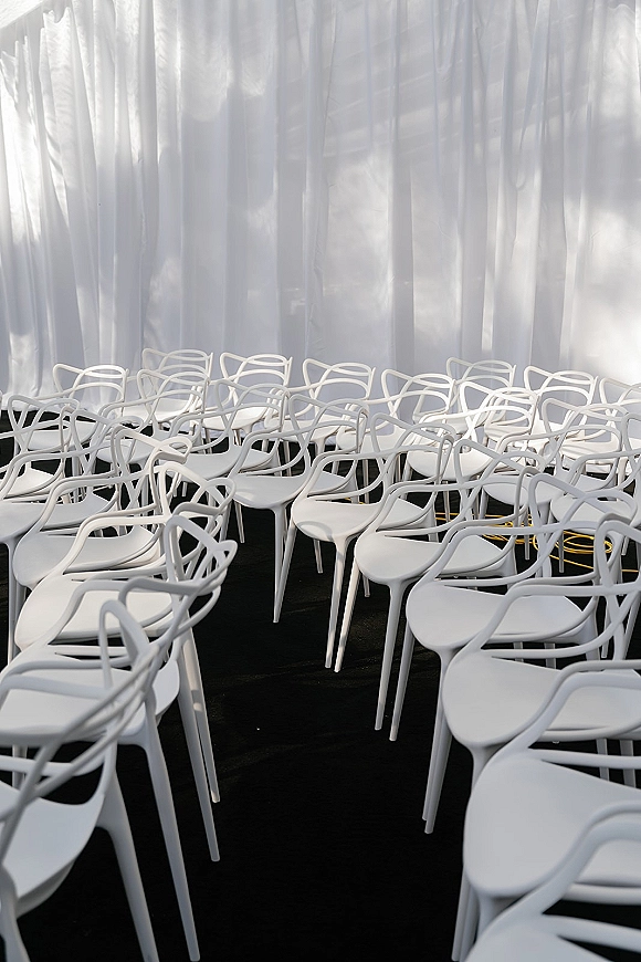 Ceremony seating with modern wedding chairs in neat white rows beside a black aisle runner, framed by sheer curtain backdrop and draped fabric indoors