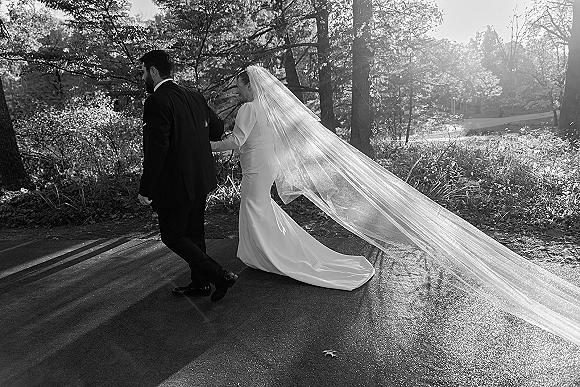 Couple portrait of bride and groom walking away, her cathedral veil train flowing in sunlight on a wooded road at golden hour