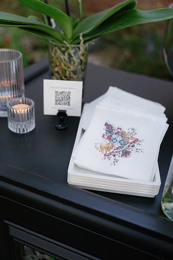 Wedding stationery with wedding crest stationery cards and QR code sign on a black tabletop, surrounded by orchids and votive candles in greenery backdrop