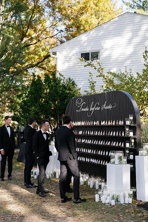 Wedding escort display featuring an escort card wall on a black backdrop with calligraphy sign, pillar candles, and white roses on an outdoor lawn