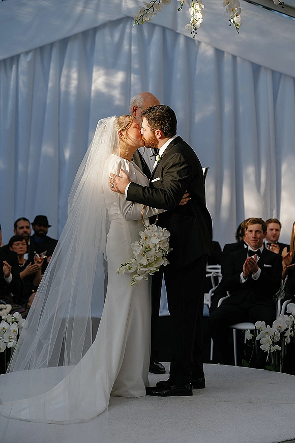 Wedding kiss as bride in long sleeve dress and veil embraces groom in tuxedo before white draped backdrop with orchids outdoors
