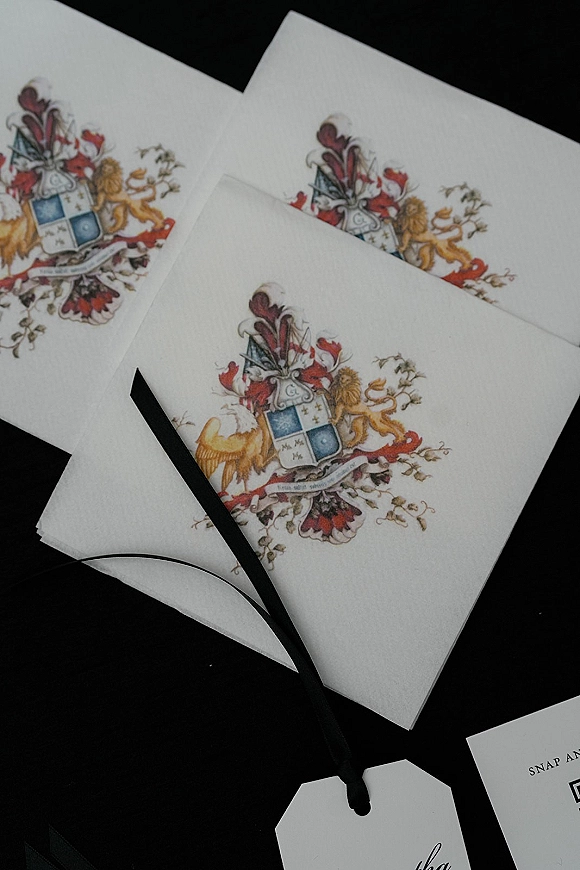 Wedding invitation suite with a custom wedding crest, envelopes, black ribbon wrap, and gift tag arranged on a black surface