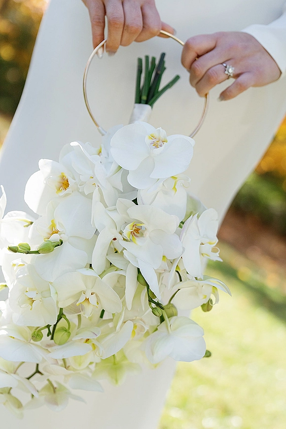 Bridal bouquet of white orchids cascading from a gold hoop handle, held in a white robe with engagement ring against sunlit garden bokeh