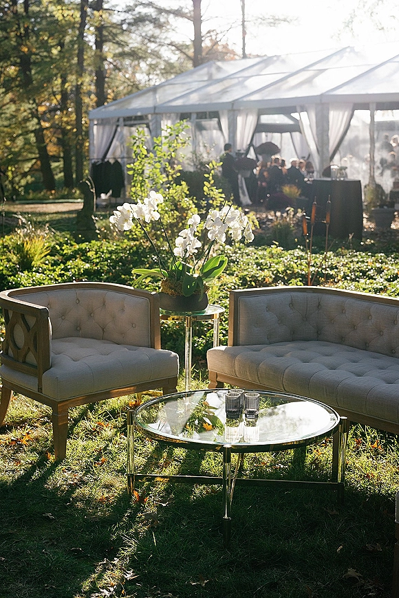 Wedding lounge area with tufted loveseat and chair around a glass coffee table, orchid centerpiece and candles under a clear tent on a sunny lawn