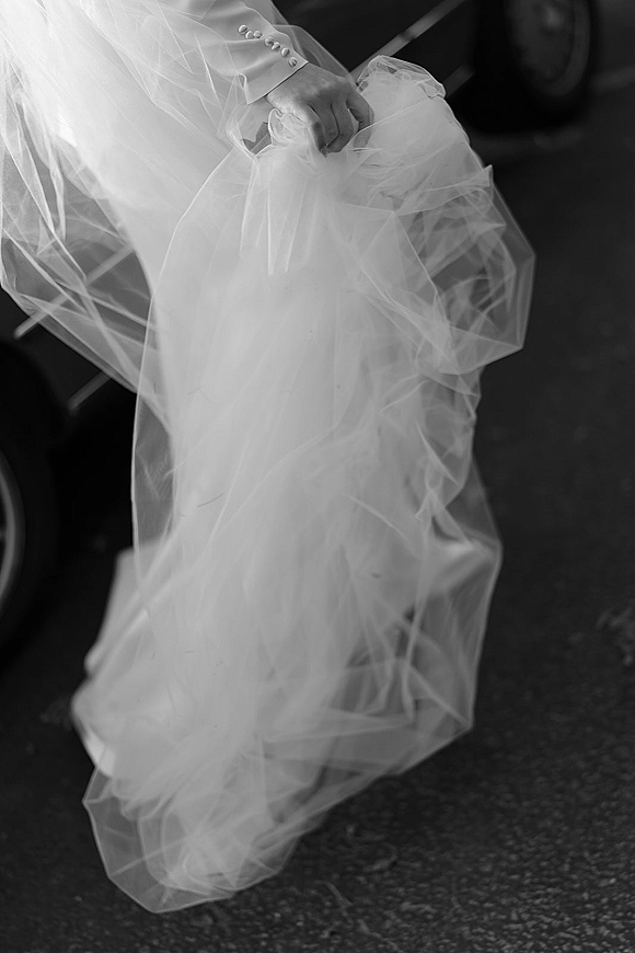 Wedding dress close-up showing tulle wedding dress texture and sleeve buttons as bride lifts hem beside dark suit trousers on asphalt ground