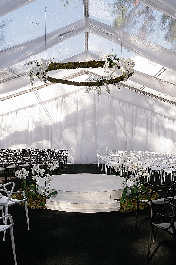 Ceremony setup in a clear top tent ceremony with round stage steps, black and white chairs, and a hanging white orchid chandelier overhead.