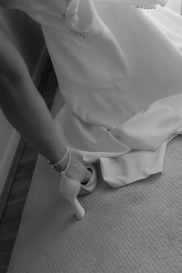 Bridal shoes, white wedding heels with ankle straps, shown beside the wedding dress hem and bracelet on a carpeted floor edge