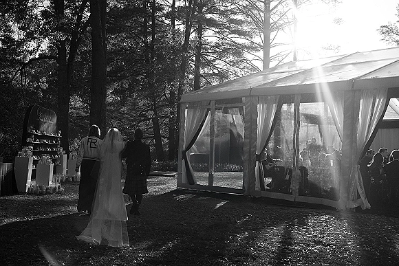 Reception entrance as bride enters with wedding veil and bridal gown toward a clear top tent, candle and escort card displays, guests in sunlight among pines