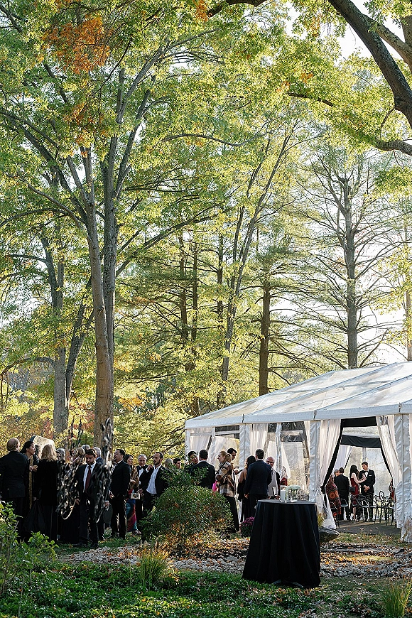 Outdoor wedding reception under a white event tent with draped curtains and black cocktail table on a stone patio amid fall trees
