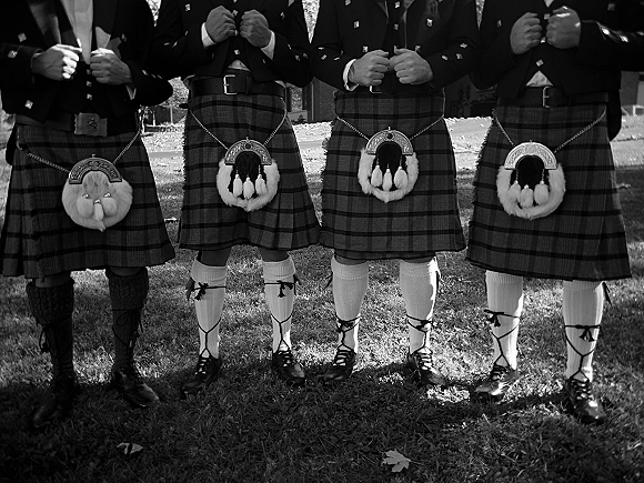Groomsmen attire featuring men in kilts with tartan kilt patterns, sporrans, and bow ties standing on a sunlit park lawn with trees