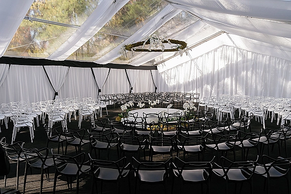 Ceremony setup in a clear top wedding tent with round black and white chair seating, white draping, and a hanging floral chandelier hoop