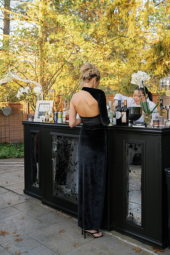 Wedding bar setup with liquor, wine and champagne bottles on a black cabinet, orchid accents and candles on an outdoor patio by trees and brick wall