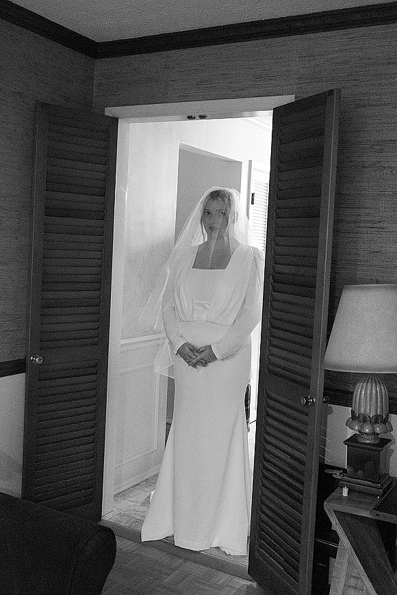 Bridal portrait in black and white of a bride in a doorway wearing a long sleeve square neckline gown and veil, with lamp-lit room behind