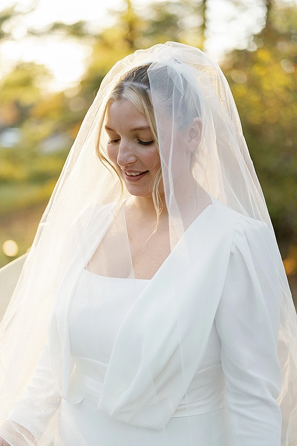 Bridal portrait of a bride wearing veil over a white bridal dress, backlit at golden hour among trees and outdoor greenery