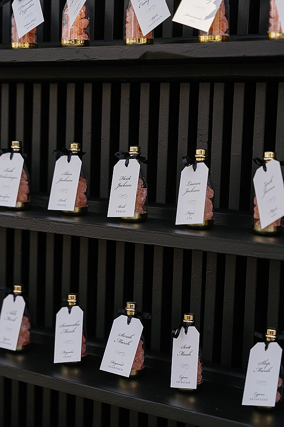 Wedding place cards as a modern escort card display with glass bottles of pink bath salts, white calligraphy tags, and black ribbon on shelves
