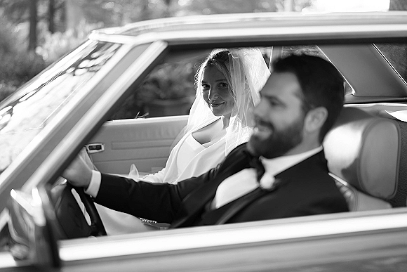 Couple portrait of bride and groom in car, her veil draped over her face as he drives a vintage getaway car with trees outside window