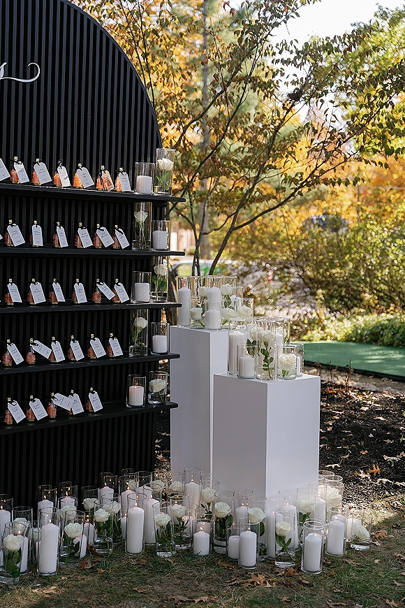 Wedding welcome display with wedding escort card display on a black arched wall, mini bottle favors, candles, and white roses in an autumn garden