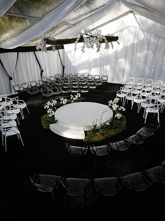 Ceremony setup with a round wedding ceremony stage surrounded by black and white chairs, white orchids, moss floral ring, and draping under a clear tent canopy
