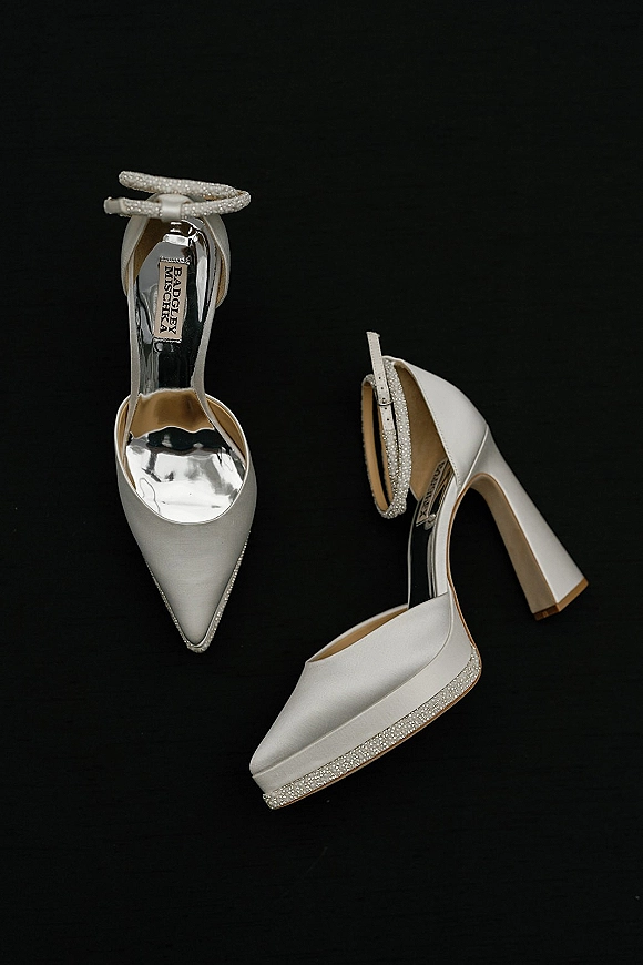 Wedding shoes styled as bridal heels with ankle strap and rhinestone detail, white platform block heels set against a black backdrop